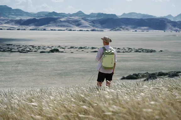 Mann mit Rucksack und Hut in der Savanne, im Hintergrund eine Gebirgskette